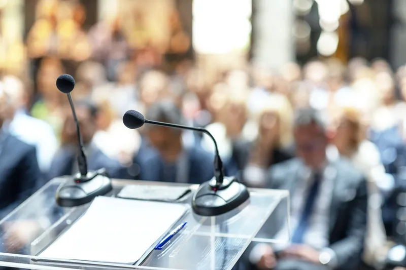 Rednerpult aus Acrylglas mit zwei Mikrofonen und Manuskript, dahinter unscharf ein voll besetzter Konferenzsaal