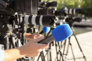 Reporterhand mit blauem Mikrofon vor professionellen Videokameras auf Stativen bei einer Pressekonferenz im Freien