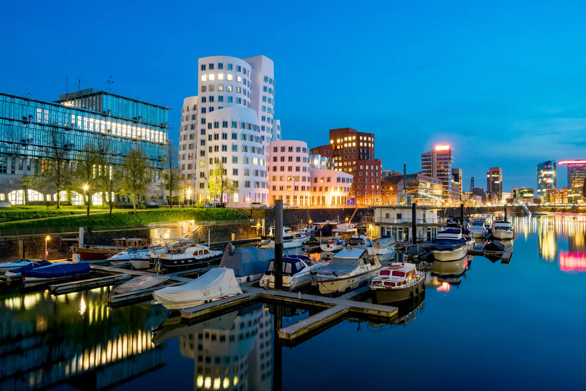 Moderne Architektur im Düsseldorfer MedienHafen mit geschwungenen Fassaden, beleuchteter Skyline und Yachten im Vordergrund.