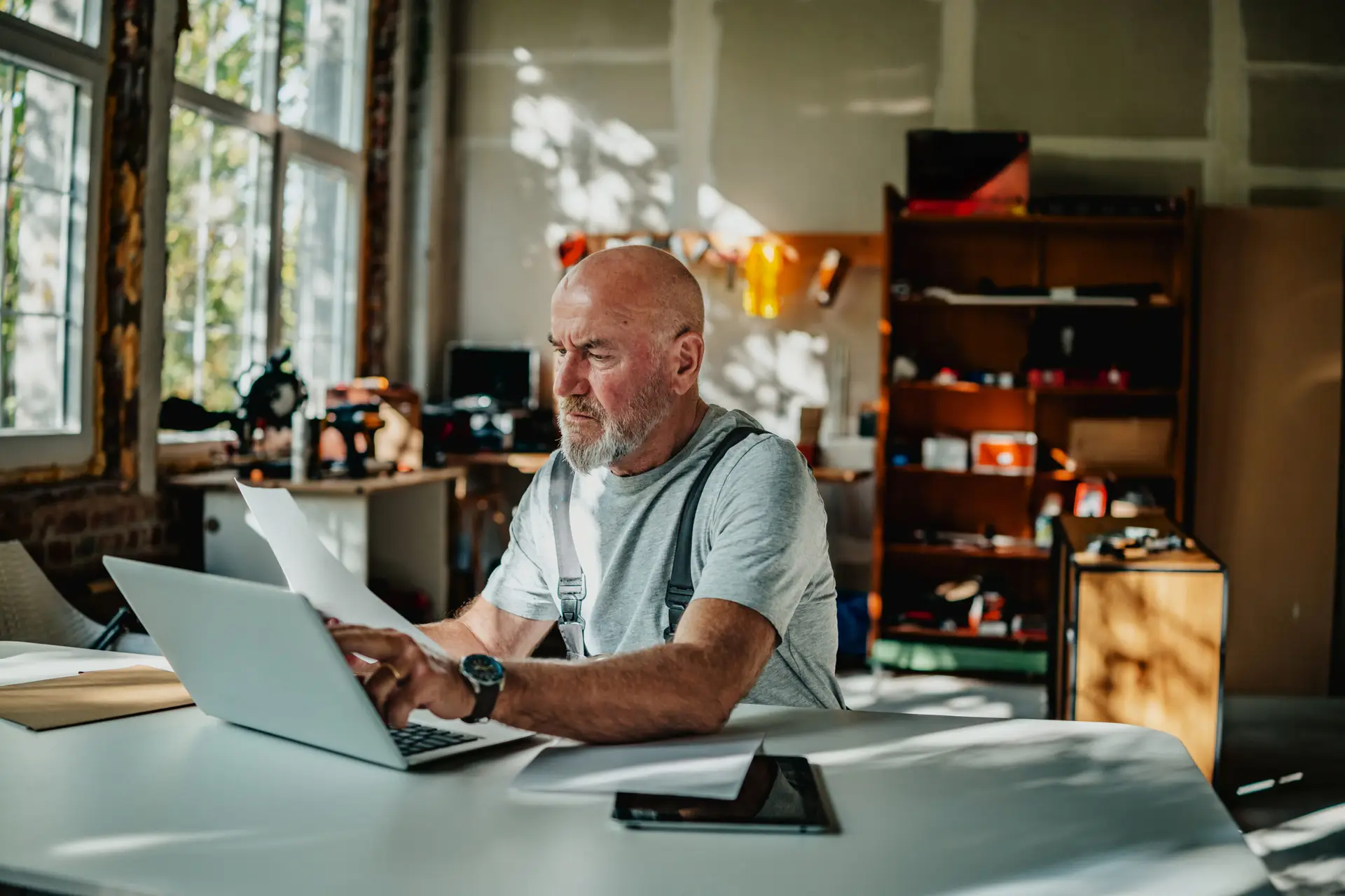 Älterer Mann arbeitet am Laptop und prüft Dokumente in Werkstatt oder Homeoffice bei Tageslicht