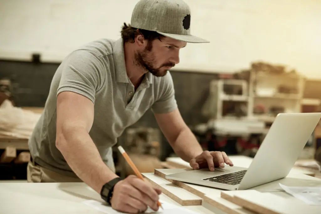 Handwerker arbeitet in einer Werkstatt mit Laptop, Bleistift und zugeschnittenen Holzplatten