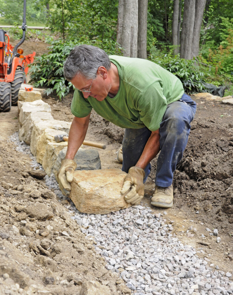 Gartenbauer setzt einen großen Naturstein in ein vorbereitetes Kiesbett für eine niedrige Trockenmauer